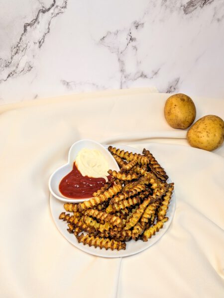 knusprige, leckere, selbstgemachte Ofen-Pommes aus dem Backofen mit Ketchup und Mayonnaise auf einem Teller serviert mit Kartoffeln im Hintergrund vor Marmor auf einem beige-farbigen Tuch