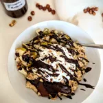 Wärmender Schoko-Nuss-Hirsebrei in einer weißen Schüssel, serviert mit frischen Birnenwürfeln, gerösteten Nüssen, geschmolzener Zartbitterschokolade und einem Klecks Skyr, dekoriert mit selbstgemachtem gesünderem Zartbitter "Nutella" im Hintergrund, auf weißem Tuch arrangiert vor Holzhintergrund.