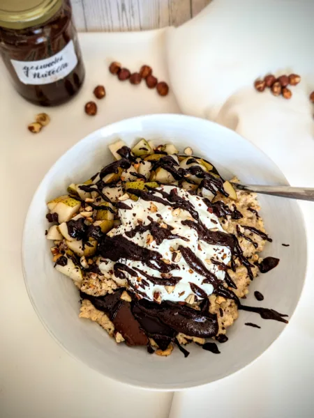 Rezepte Wärmender Schoko-Nuss-Hirsebrei in einer weißen Schüssel, serviert mit frischen Birnenwürfeln, gerösteten Nüssen, geschmolzener Zartbitterschokolade und einem Klecks Skyr, dekoriert mit selbstgemachtem gesünderem Zartbitter "Nutella" im Hintergrund, auf weißem Tuch arrangiert vor Holzhintergrund.