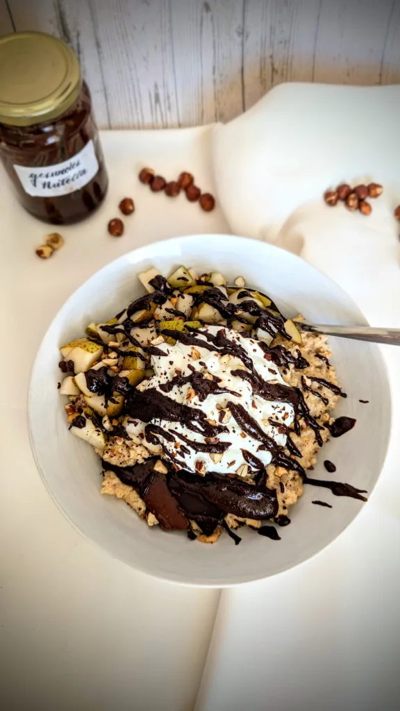 Chocolatey & Nutty Millet Porridge with Pear and Cinnamon Wärmender Schoko-Nuss-Hirsebrei in einer weißen Schüssel, serviert mit frischen Birnenwürfeln, gerösteten Nüssen, geschmolzener Zartbitterschokolade und einem Klecks Skyr, dekoriert mit selbstgemachtem gesünderem Zartbitter "Nutella" im Hintergrund, auf weißem Tuch arrangiert vor Holzhintergrund.