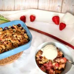 Ein Schälchen mit warmem Erdbeer-Rhabarber-Crumble, serviert mit einer Kugel cremigem Vanilleeis. Im Hintergrund eine Auflaufform mit dem restlichen Crumble sowie frische Erdbeeren und ein Rhabarberstiel. Das fruchtige Dessert ist auf einem hellen Untergrund angerichtet. Sommerliches Erdbeer-Rhabarber-Crumble mit Vanilleeis – einfaches Dessert-Rezept mit einem Hauch von Limette.