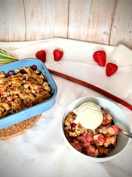 Rezepte Ein Schälchen mit warmem Erdbeer-Rhabarber-Crumble, serviert mit einer Kugel cremigem Vanilleeis. Im Hintergrund eine Auflaufform mit dem restlichen Crumble sowie frische Erdbeeren und ein Rhabarberstiel. Das fruchtige Dessert ist auf einem hellen Untergrund angerichtet. Sommerliches Erdbeer-Rhabarber-Crumble mit Vanilleeis – einfaches Dessert-Rezept mit einem Hauch von Limette.