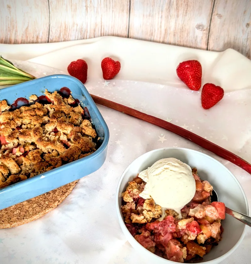 Ein Schälchen mit warmem Erdbeer-Rhabarber-Crumble, serviert mit einer Kugel cremigem Vanilleeis. Im Hintergrund eine Auflaufform mit dem restlichen Crumble sowie frische Erdbeeren und ein Rhabarberstiel. Das fruchtige Dessert ist auf einem hellen Untergrund angerichtet. Sommerliches Erdbeer-Rhabarber-Crumble mit Vanilleeis – einfaches Dessert-Rezept mit einem Hauch von Limette.