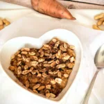 Herzförmige Schale mit cremigem Süßkartoffel-Erdnuss-Porridge, getoppt mit Erdnüssen und Zimt, angerichtet auf weißem Tuch mit Löffel daneben. Im Hintergrund liegt eine Süßkartoffel und einige Erdnüsse auf rustikalem Holzuntergrund.