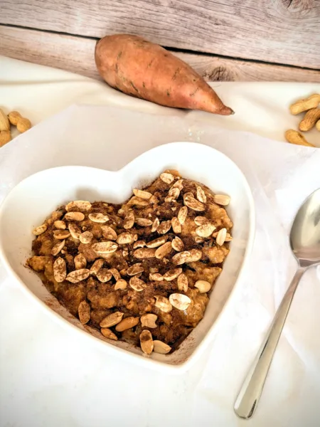 Herzförmige Schale mit cremigem Süßkartoffel-Erdnuss-Porridge, getoppt mit Erdnüssen und Zimt, angerichtet auf weißem Tuch mit Löffel daneben. Im Hintergrund liegt eine Süßkartoffel und einige Erdnüsse auf rustikalem Holzuntergrund.