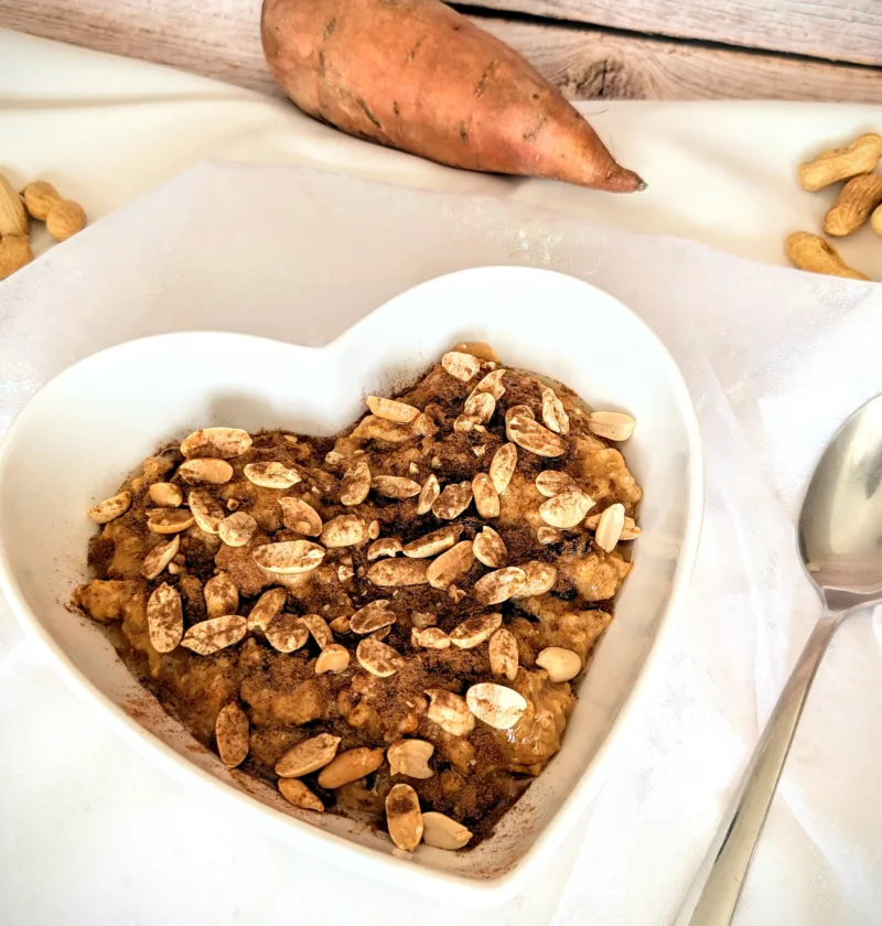 Herzförmige Schale mit cremigem Süßkartoffel-Erdnuss-Porridge, getoppt mit Erdnüssen und Zimt, angerichtet auf weißem Tuch mit Löffel daneben. Im Hintergrund liegt eine Süßkartoffel und einige Erdnüsse auf rustikalem Holzuntergrund.