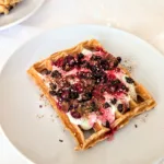 Frisch gebackene Quarkwaffeln aus Dinkelvollkornmehl auf einem weißen Teller, serviert mit Joghurt, roten Beeren, gepufftem Quinoa und Zimt – gesundes Waffelrezept für ein ausgewogenes Frühstück oder Brunch.
