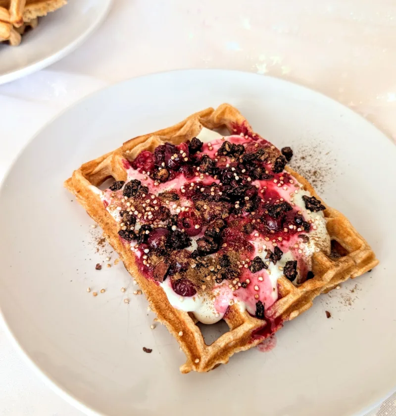 Frisch gebackene Quarkwaffeln aus Dinkelvollkornmehl auf einem weißen Teller, serviert mit Joghurt, roten Beeren, gepufftem Quinoa und Zimt – gesundes Waffelrezept für ein ausgewogenes Frühstück oder Brunch.
