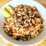 Authentisches Pad Thai mit Reisnudeln, frischem Tofu, Erdnüssen, Limette und Frühlingszwiebeln – auf einem Teller angerichtet, typisch thailändisch und bunt, perfekt für die Follikelphase geeignet. Der Teller ist auf einem weißen und goldfarbigen Tuch angerichtet mit einem holzfarbigen Hintergrund.