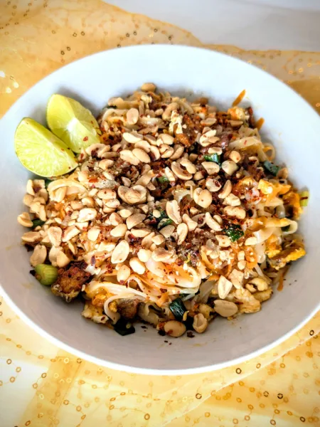 Pad Thai Authentisches Pad Thai mit Reisnudeln, frischem Tofu, Erdnüssen, Limette und Frühlingszwiebeln – auf einem Teller angerichtet, typisch thailändisch und bunt, perfekt für die Follikelphase geeignet. Der Teller ist auf einem weißen und goldfarbigen Tuch angerichtet mit einem holzfarbigen Hintergrund.