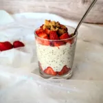 Ein Glas mit cremigen Strawberry-Cheesecake-Overnight-Oats, geschichtet mit frischen Erdbeerstücken und getoppt mit Butterkeks-Krümeln. Das Frühstück steht auf einem hellen, mit Glitzersternen bestickten Stoff, im Hintergrund eine Holzoptik. Die Erdbeeren am Boden des Glases sind dekorativ am Rand platziert und sorgen für ein ansprechendes, sommerliches Aussehen.