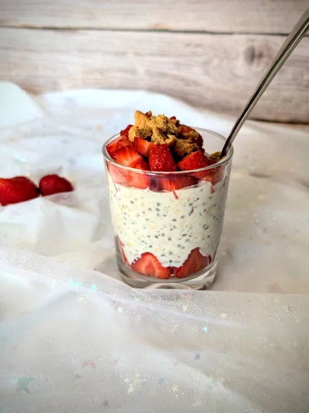 Strawberry-Cheesecake-Overnight-Oats Ein Glas mit cremigen Strawberry-Cheesecake-Overnight-Oats, geschichtet mit frischen Erdbeerstücken und getoppt mit Butterkeks-Krümeln. Das Frühstück steht auf einem hellen, mit Glitzersternen bestickten Stoff, im Hintergrund eine Holzoptik. Die Erdbeeren am Boden des Glases sind dekorativ am Rand platziert und sorgen für ein ansprechendes, sommerliches Aussehen.