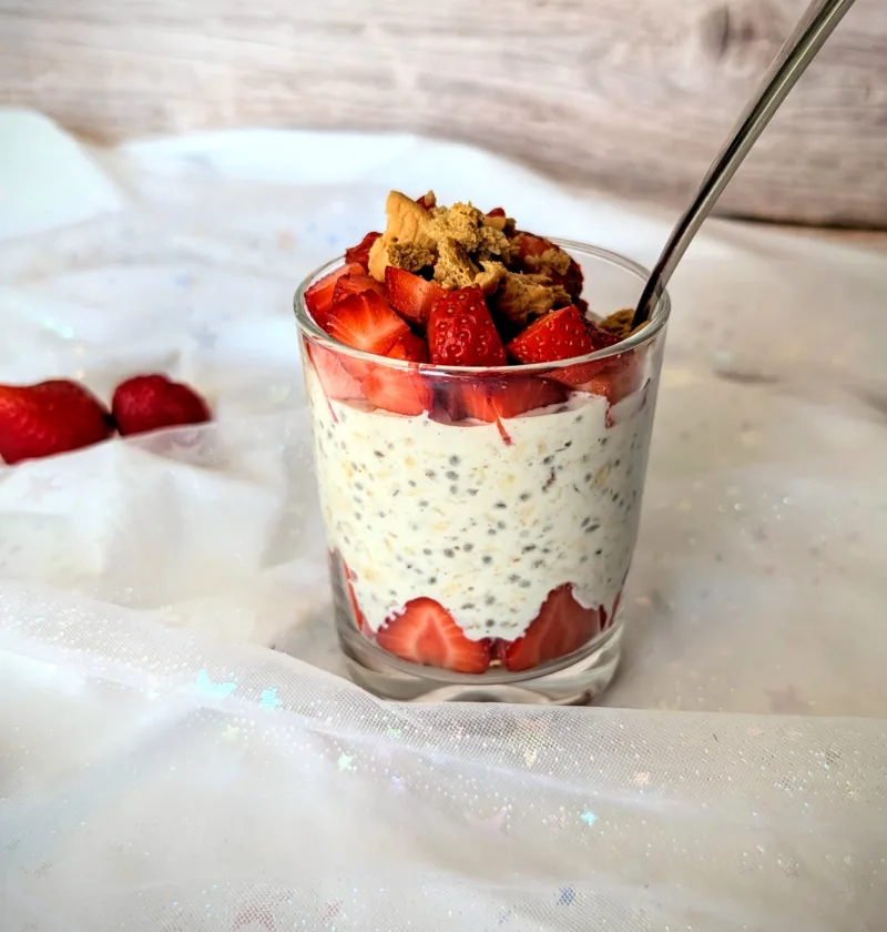 Ein Glas mit cremigen Strawberry-Cheesecake-Overnight-Oats, geschichtet mit frischen Erdbeerstücken und getoppt mit Butterkeks-Krümeln. Das Frühstück steht auf einem hellen, mit Glitzersternen bestickten Stoff, im Hintergrund eine Holzoptik. Die Erdbeeren am Boden des Glases sind dekorativ am Rand platziert und sorgen für ein ansprechendes, sommerliches Aussehen.