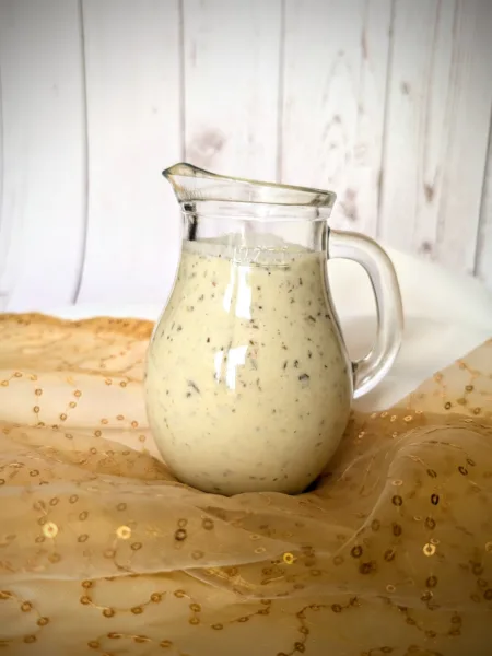 Recipes Ein kleines, bauchiges Glasgefäß mit cremigem Kräuter-Joghurt-Dressing mit sichtbaren Kräuterstückchen. Das Gefäß steht auf einem goldfarbenen, leicht transparenten Tuch mit Pailletten. Im Hintergrund ist eine weiße, vertäfelte Holzfläche zu sehen.