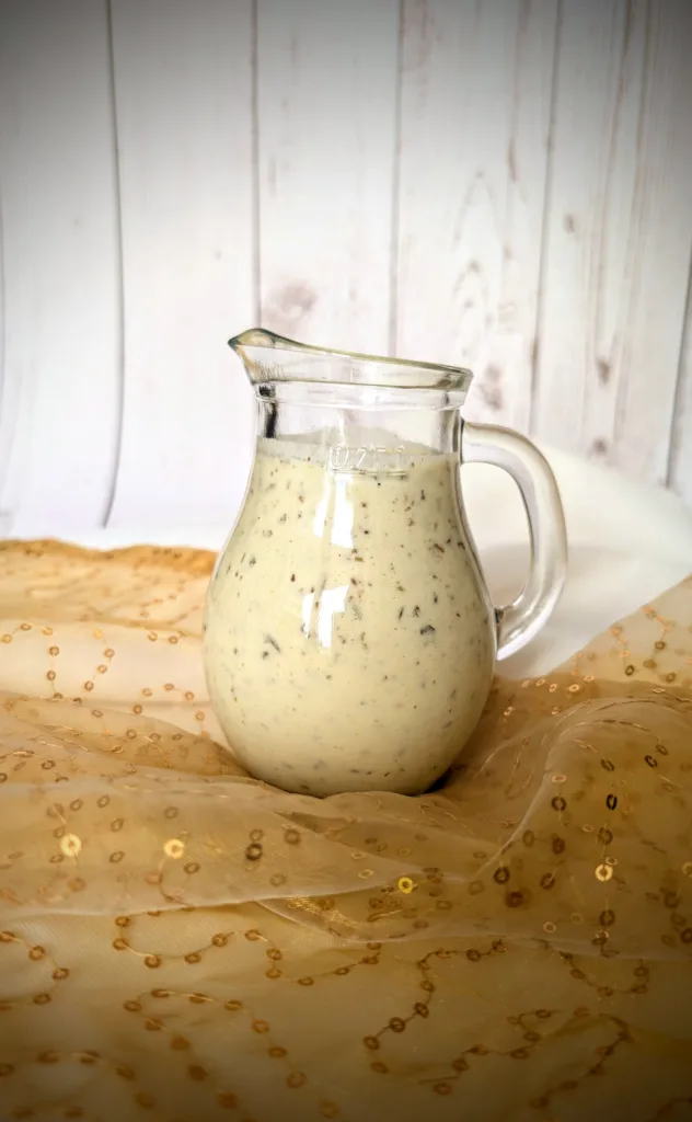 Kräuter-Joghurt-Dressing Ein kleines, bauchiges Glasgefäß mit cremigem Kräuter-Joghurt-Dressing mit sichtbaren Kräuterstückchen. Das Gefäß steht auf einem goldfarbenen, leicht transparenten Tuch mit Pailletten. Im Hintergrund ist eine weiße, vertäfelte Holzfläche zu sehen.
