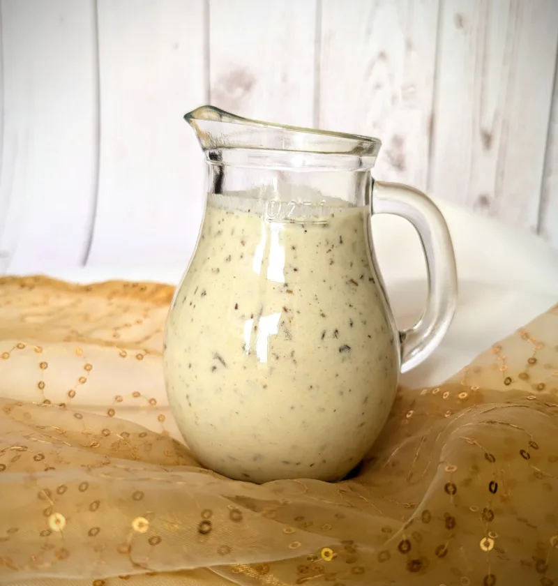 Ein kleines, bauchiges Glasgefäß mit cremigem Kräuter-Joghurt-Dressing mit sichtbaren Kräuterstückchen. Das Gefäß steht auf einem goldfarbenen, leicht transparenten Tuch mit Pailletten. Im Hintergrund ist eine weiße, vertäfelte Holzfläche zu sehen.
