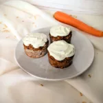 Drei Karottenkuchen-Muffins mit Cream-Cheese-Frosting auf einem weißen Teller, dekoriert mit Haferflocken und einer frischen Karotte im Hintergrund – gesundes Frühstücksrezept mit Rosinen und Walnüssen, super geeignet für die Follikelphase.