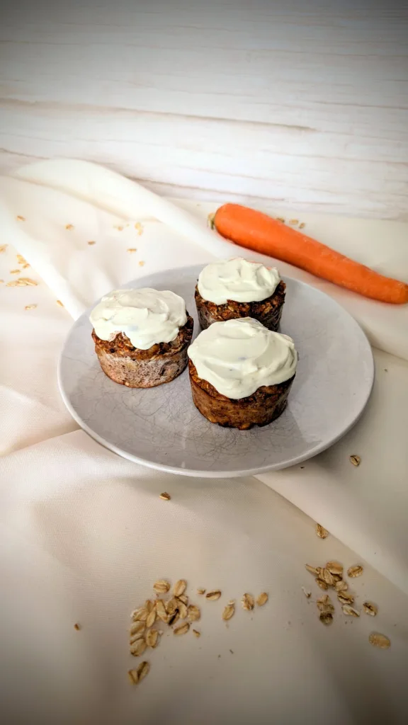 Karottenkuchen-Muffins mit Haferflocken Drei Karottenkuchen-Muffins mit Cream-Cheese-Frosting auf einem weißen Teller, dekoriert mit Haferflocken und einer frischen Karotte im Hintergrund – gesundes Frühstücksrezept mit Rosinen und Walnüssen, super geeignet für die Follikelphase.