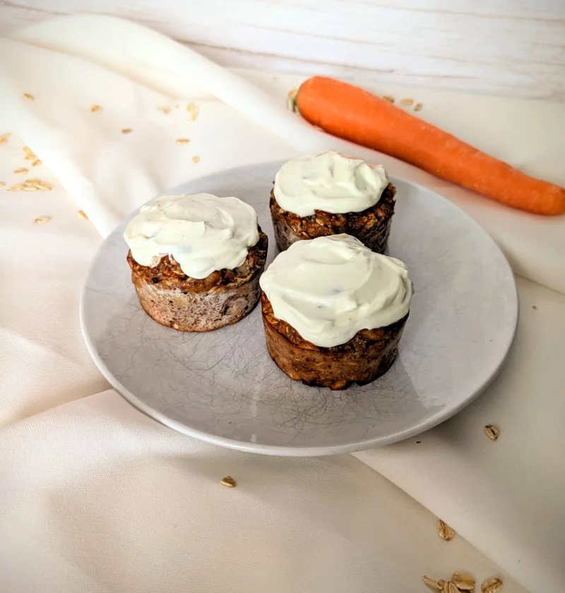 Drei Karottenkuchen-Muffins mit Cream-Cheese-Frosting auf einem weißen Teller, dekoriert mit Haferflocken und einer frischen Karotte im Hintergrund – gesundes Frühstücksrezept mit Rosinen und Walnüssen, super geeignet für die Follikelphase.
