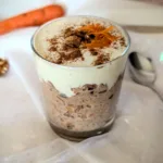 Ein dekorativ angerichtetes Glas mit Karottenkuchen-Porridge, getoppt mit Cream-Cheese-Frosting, Zimt, Walnüssen und frischen Karottenraspeln. Daneben liegt ein Löffel auf einem zarten, durchsichtigen Stoff mit weißen Sternen. Das Bild zeigt ein kreatives, gesundes Frühstücksrezept in ästhetischer Food-Inszenierung.