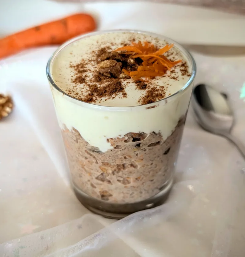 Ein dekorativ angerichtetes Glas mit Karottenkuchen-Porridge, getoppt mit Cream-Cheese-Frosting, Zimt, Walnüssen und frischen Karottenraspeln. Daneben liegt ein Löffel auf einem zarten, durchsichtigen Stoff mit weißen Sternen. Das Bild zeigt ein kreatives, gesundes Frühstücksrezept in ästhetischer Food-Inszenierung.