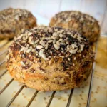 Nahaufnahme eines 100 % Weizenvollkorn Körner Burger Buns mit Sauerteig, üppig bestreut mit Sonnenblumenkernen, Sesam, Mohn und Leinsamen, auf einem Kuchengitter mit golden gemustertem Stoff darunter.
