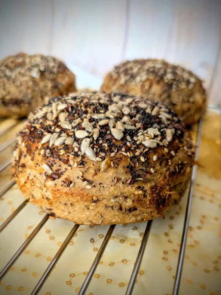 Recipes Nahaufnahme eines 100 % Weizenvollkorn Körner Burger Buns mit Sauerteig, üppig bestreut mit Sonnenblumenkernen, Sesam, Mohn und Leinsamen, auf einem Kuchengitter mit golden gemustertem Stoff darunter.