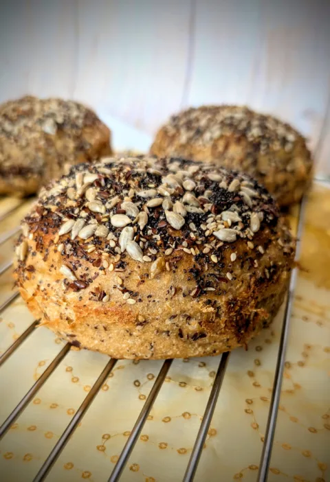 Nahaufnahme eines 100 % Weizenvollkorn Körner Burger Buns mit Sauerteig, üppig bestreut mit Sonnenblumenkernen, Sesam, Mohn und Leinsamen, auf einem Kuchengitter mit golden gemustertem Stoff darunter.