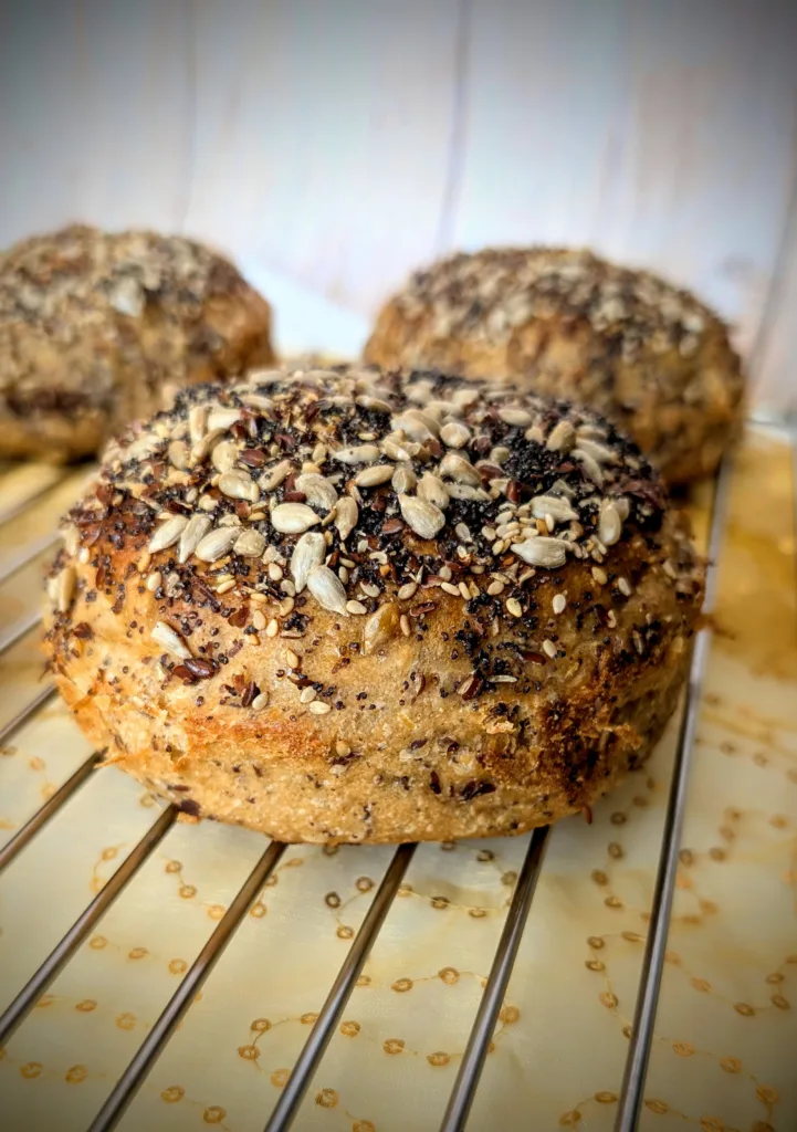 Fluffy 100 % Wholewheat Seeded Burger Buns with Sourdough Nahaufnahme eines 100 % Weizenvollkorn Körner Burger Buns mit Sauerteig, üppig bestreut mit Sonnenblumenkernen, Sesam, Mohn und Leinsamen, auf einem Kuchengitter mit golden gemustertem Stoff darunter.