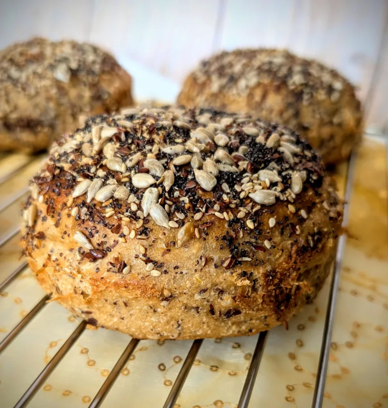 Nahaufnahme eines 100 % Weizenvollkorn Körner Burger Buns mit Sauerteig, üppig bestreut mit Sonnenblumenkernen, Sesam, Mohn und Leinsamen, auf einem Kuchengitter mit golden gemustertem Stoff darunter.