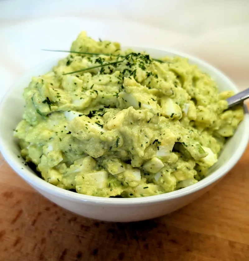 Cremiger Avocado-Eiersalat in einer weißen Schale, garniert mit frischen Kräutern, serviert auf einem Holzbrett – proteinreiches und hormonfreundliches Rezept für die Follikelphase.