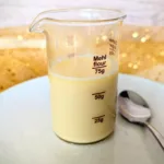 Glasbecher mit selbstgemachter zuckerfreier gesüßter Kondensmilch, zubereitet mit Erythrit, auf einem weißen Teller mit Löffel. Perfekt als Low-Carb-Basis für Desserts, Pumpkin Spice Latte oder Eisrezepte.