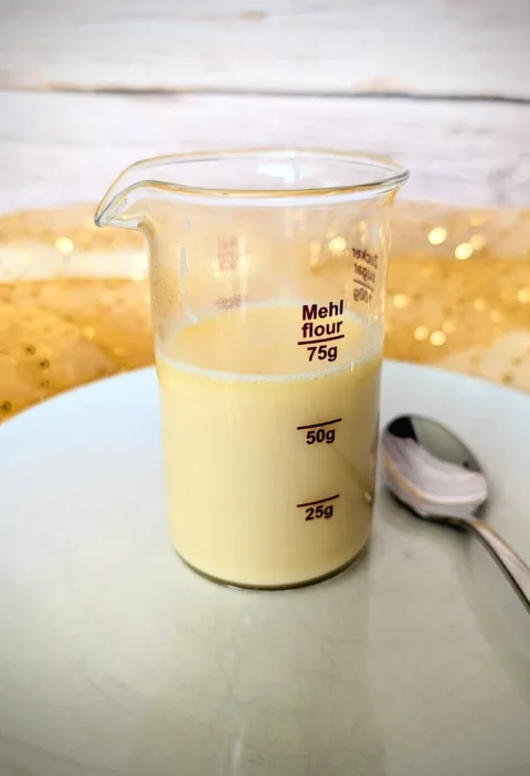 Glasbecher mit selbstgemachter zuckerfreier gesüßter Kondensmilch, zubereitet mit Erythrit, auf einem weißen Teller mit Löffel. Perfekt als Low-Carb-Basis für Desserts, Pumpkin Spice Latte oder Eisrezepte.