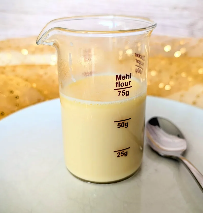 Glasbecher mit selbstgemachter zuckerfreier gesüßter Kondensmilch, zubereitet mit Erythrit, auf einem weißen Teller mit Löffel. Perfekt als Low-Carb-Basis für Desserts, Pumpkin Spice Latte oder Eisrezepte.