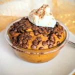 Kürbis Chocolate Chip Baked Oats in einer kleinen Glasschüssel, goldbraun gebacken und mit vielen geschmolzenen Schokostückchen bedeckt. Obenauf ein Klecks cremiger Skyr, bestäubt mit Zimt. Das herbstliche Frühstück steht auf einem weißen Teller vor goldenem, leicht glänzendem Stoffhintergrund.