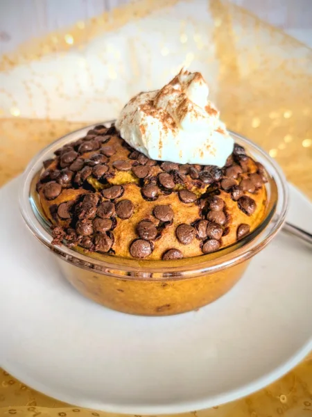 Kürbis Chocolate Chip Baked Oats in einer kleinen Glasschüssel, goldbraun gebacken und mit vielen geschmolzenen Schokostückchen bedeckt. Obenauf ein Klecks cremiger Skyr, bestäubt mit Zimt. Das herbstliche Frühstück steht auf einem weißen Teller vor goldenem, leicht glänzendem Stoffhintergrund.