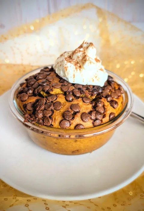 Kürbis Chocolate Chip Baked Oats in einer kleinen Glasschüssel, goldbraun gebacken und mit vielen geschmolzenen Schokostückchen bedeckt. Obenauf ein Klecks cremiger Skyr, bestäubt mit Zimt. Das herbstliche Frühstück steht auf einem weißen Teller vor goldenem, leicht glänzendem Stoffhintergrund.