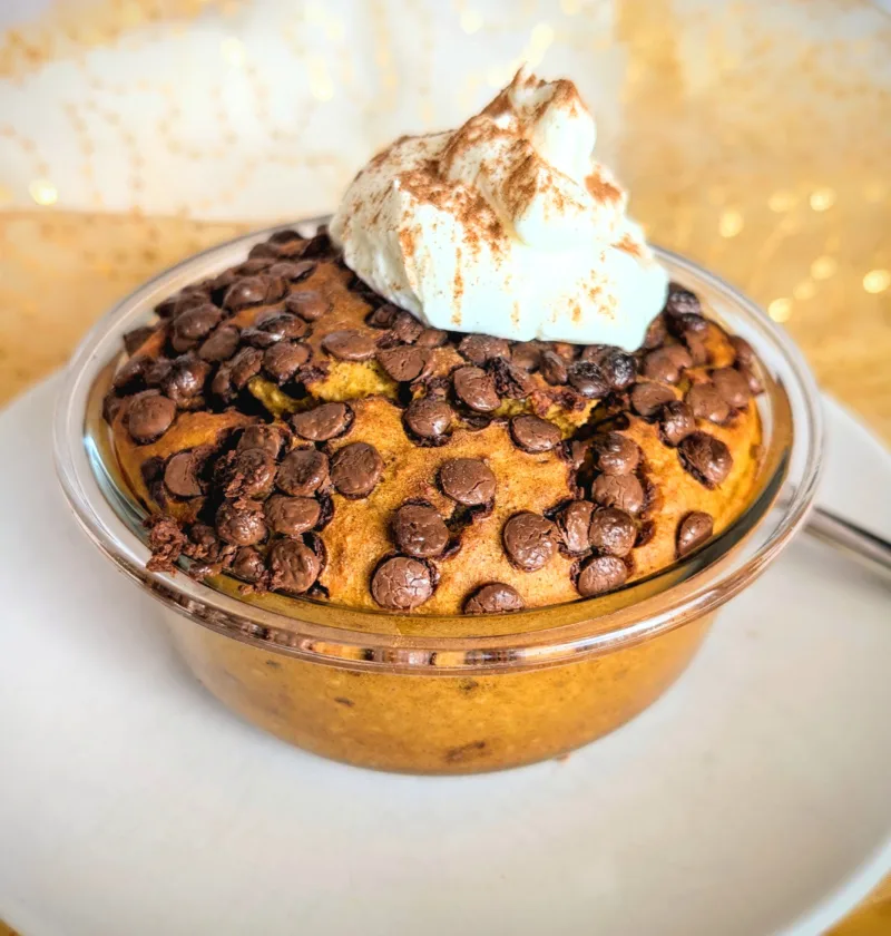 Kürbis Chocolate Chip Baked Oats in einer kleinen Glasschüssel, goldbraun gebacken und mit vielen geschmolzenen Schokostückchen bedeckt. Obenauf ein Klecks cremiger Skyr, bestäubt mit Zimt. Das herbstliche Frühstück steht auf einem weißen Teller vor goldenem, leicht glänzendem Stoffhintergrund.