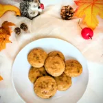 Ein Teller mit mehreren „Dirty Chai Cookies“ (von Taylor Swift inspiriert), glasiert mit brauner Butter-Zuckerguss, steht auf einem hellen Tuch. Um den Teller herum liegen herbstliche Dekorationen wie bunte Blätter, Zapfen, kleine Kürbisse und eine Igel-Figur. Das Bild vermittelt eine gemütliche Herbststimmung.