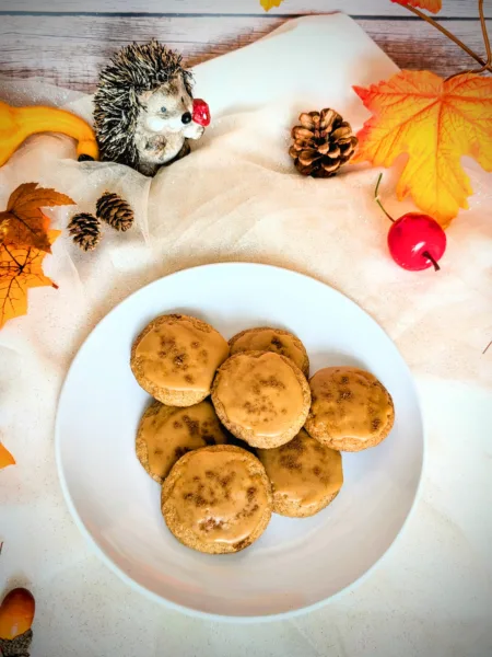 Taylor Swift inspired Dirty Chai Cookies Ein Teller mit mehreren „Dirty Chai Cookies“ (von Taylor Swift inspiriert), glasiert mit brauner Butter-Zuckerguss, steht auf einem hellen Tuch. Um den Teller herum liegen herbstliche Dekorationen wie bunte Blätter, Zapfen, kleine Kürbisse und eine Igel-Figur. Das Bild vermittelt eine gemütliche Herbststimmung.