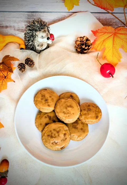 Ein Teller mit mehreren „Dirty Chai Cookies“ (von Taylor Swift inspiriert), glasiert mit brauner Butter-Zuckerguss, steht auf einem hellen Tuch. Um den Teller herum liegen herbstliche Dekorationen wie bunte Blätter, Zapfen, kleine Kürbisse und eine Igel-Figur. Das Bild vermittelt eine gemütliche Herbststimmung.