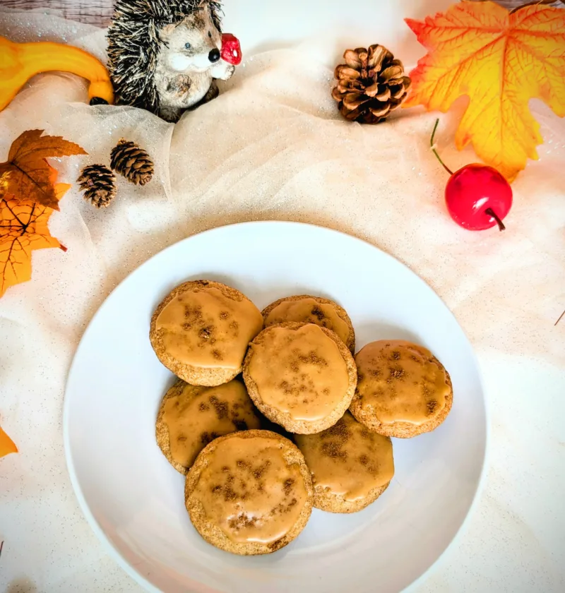 Ein Teller mit mehreren „Dirty Chai Cookies“ (von Taylor Swift inspiriert), glasiert mit brauner Butter-Zuckerguss, steht auf einem hellen Tuch. Um den Teller herum liegen herbstliche Dekorationen wie bunte Blätter, Zapfen, kleine Kürbisse und eine Igel-Figur. Das Bild vermittelt eine gemütliche Herbststimmung.