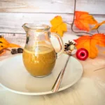 Ein Glas mit cremigem, selbstgemachtem Pumpkin Spice Sirup steht auf einem weißen Teller mit Löffel, umgeben von herbstlicher Deko in warmen Orange- und Gelbtönen. Der zuckerfreie Sirup nach Starbucks-Vorbild eignet sich perfekt für Pumpkin Spice Latte und verleiht Getränken einen aromatisch-würzigen Geschmack.