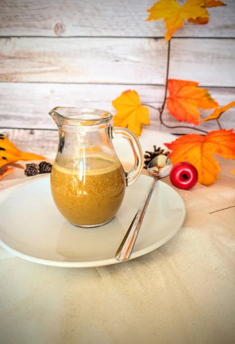 Ein Glas mit cremigem, selbstgemachtem Pumpkin Spice Sirup steht auf einem weißen Teller mit Löffel, umgeben von herbstlicher Deko in warmen Orange- und Gelbtönen. Der zuckerfreie Sirup nach Starbucks-Vorbild eignet sich perfekt für Pumpkin Spice Latte und verleiht Getränken einen aromatisch-würzigen Geschmack.