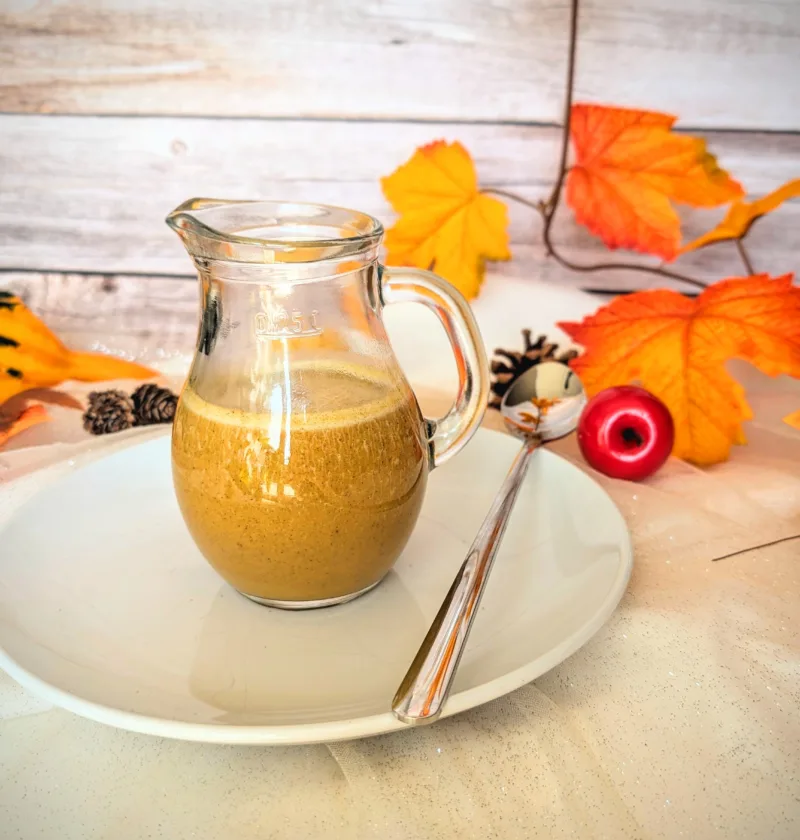Ein Glas mit cremigem, selbstgemachtem Pumpkin Spice Sirup steht auf einem weißen Teller mit Löffel, umgeben von herbstlicher Deko in warmen Orange- und Gelbtönen. Der zuckerfreie Sirup nach Starbucks-Vorbild eignet sich perfekt für Pumpkin Spice Latte und verleiht Getränken einen aromatisch-würzigen Geschmack.