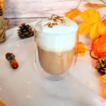 Ein zuckerfreier Pumpkin Spice Latte in einem doppelwandigen Glas, getoppt mit cremigem Milchschaum und etwas Pumpkin Spice, steht auf einer herbstlich dekorierten Oberfläche mit bunten Blättern, Tannenzapfen und einem kleinen Deko-Apfel. Dieses selbstgemachte Low-Carb-Getränk ist eine aromatische, preiswerte Alternative zum Starbucks Pumpkin Spice Latte und bringt herbstlichen Genuss direkt nach Hause.