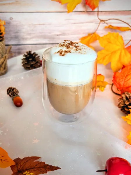 Rezepte Ein zuckerfreier Pumpkin Spice Latte in einem doppelwandigen Glas, getoppt mit cremigem Milchschaum und etwas Pumpkin Spice, steht auf einer herbstlich dekorierten Oberfläche mit bunten Blättern, Tannenzapfen und einem kleinen Deko-Apfel. Dieses selbstgemachte Low-Carb-Getränk ist eine aromatische, preiswerte Alternative zum Starbucks Pumpkin Spice Latte und bringt herbstlichen Genuss direkt nach Hause.