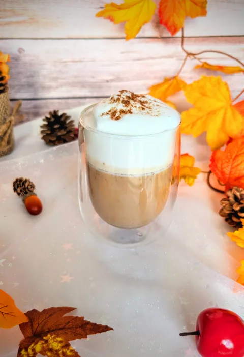 Ein zuckerfreier Pumpkin Spice Latte in einem doppelwandigen Glas, getoppt mit cremigem Milchschaum und etwas Pumpkin Spice, steht auf einer herbstlich dekorierten Oberfläche mit bunten Blättern, Tannenzapfen und einem kleinen Deko-Apfel. Dieses selbstgemachte Low-Carb-Getränk ist eine aromatische, preiswerte Alternative zum Starbucks Pumpkin Spice Latte und bringt herbstlichen Genuss direkt nach Hause.