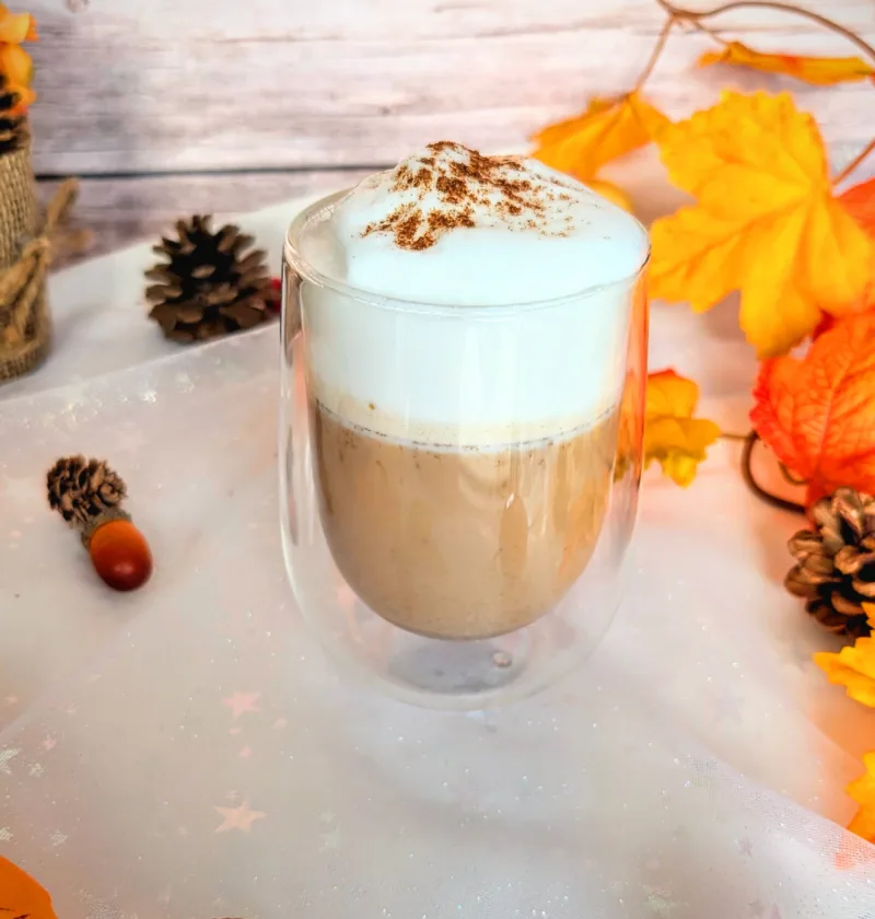 Ein zuckerfreier Pumpkin Spice Latte in einem doppelwandigen Glas, getoppt mit cremigem Milchschaum und etwas Pumpkin Spice, steht auf einer herbstlich dekorierten Oberfläche mit bunten Blättern, Tannenzapfen und einem kleinen Deko-Apfel. Dieses selbstgemachte Low-Carb-Getränk ist eine aromatische, preiswerte Alternative zum Starbucks Pumpkin Spice Latte und bringt herbstlichen Genuss direkt nach Hause.