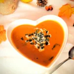 Asiatisch inspirierte Kürbissuppe mit Möhren, Süßkartoffel und Kokosmilch in einer herzförmigen weißen Schüssel, garniert mit Kürbiskernen und frischen Kräutern. Im Hintergrund herbstliche Deko mit Kürbis, Tannenzapfen und bunten Blättern.